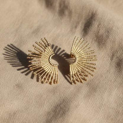 Solea Sonnenohrringe in Gold auf hellem Leinenstoff mit warmem Licht und Schatten.