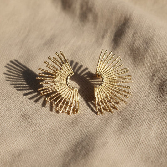 Solea Sonnenohrringe in Gold auf hellem Leinenstoff mit warmem Licht und Schatten.
