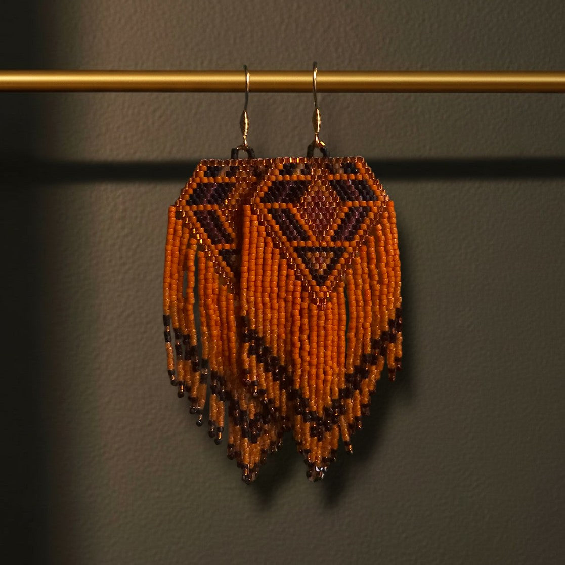 Handgewebte lange Perlenohrringe Terra Calda an einer goldenen Präsentationsstange vor einem dunklen Hintergrund. Das Design zeigt ein kraftvolles Rautenmuster in tiefem Rostbraun, warmem Orange und Schwarz mit fließenden Fransen.
