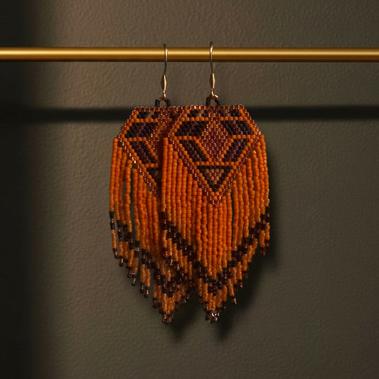 Handgewebte lange Perlenohrringe Terra Calda an einer goldenen Präsentationsstange vor einem dunklen Hintergrund. Das Design zeigt ein kraftvolles Rautenmuster in tiefem Rostbraun, warmem Orange und Schwarz mit fließenden Fransen.