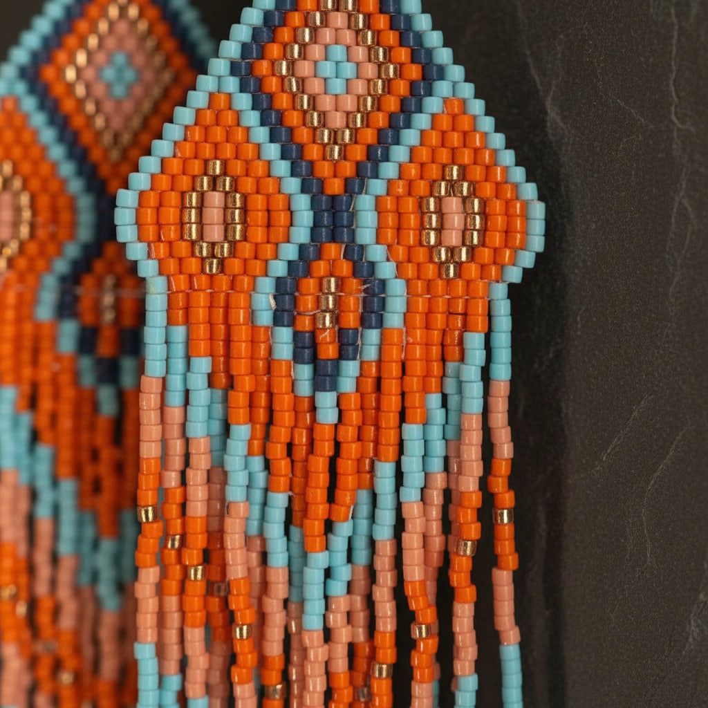 Nahaufnahme eines handgewebten Statement-Ohrrings von EDELATELIER Modell Terra e Mare. Zu sehen ist das detaillierte geometrische Rautenmuster aus hochwertigen Miyuki Delica Perlen in den Farben Terrakotta, Orange, Blau und Gold auf einem dunklen Schieferhintergrund.