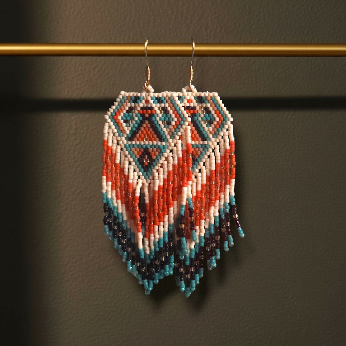 Handgewebte lange Perlenohrringe Tramonto Egeo, aufgehängt an einer goldenen Präsentationsstange vor einem dunklen Hintergrund. Das Design zeigt ein grafisches Rautenmuster in kontrastreichen Farben wie Dunkelblau, Korallenrot und Weiß mit fließenden Fransen.