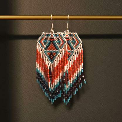 Handgewebte lange Perlenohrringe Tramonto Egeo, aufgehängt an einer goldenen Präsentationsstange vor einem dunklen Hintergrund. Das Design zeigt ein grafisches Rautenmuster in kontrastreichen Farben wie Dunkelblau, Korallenrot und Weiß mit fließenden Fransen.