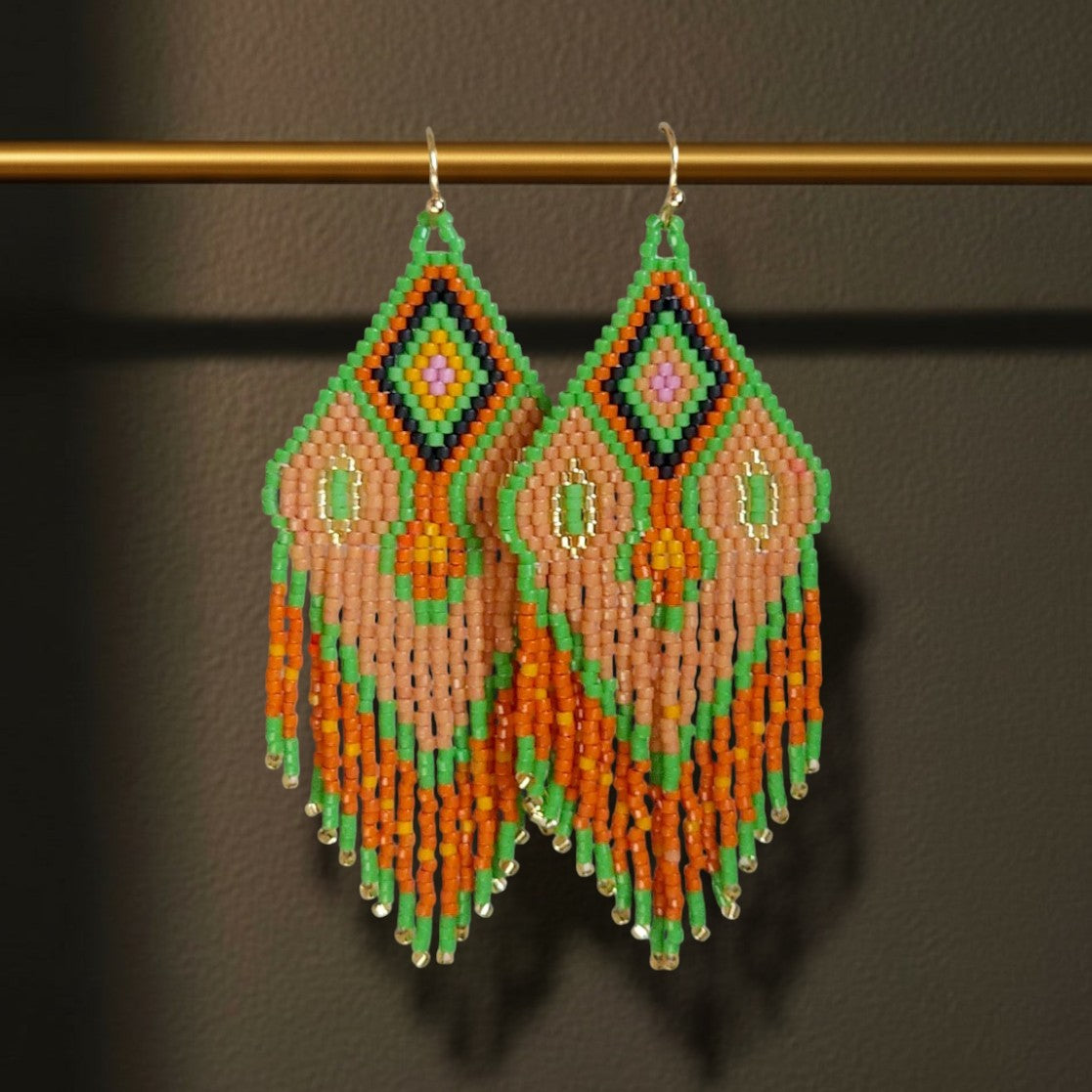 Handgewebte Miyuki Perlenohrringe Modell Tramonto Etrusco von EDELATELIER., dekorativ hängend an einer horizontalen goldenen Metallstange. Das geometrische Muster in Blattgrün, Terrakotta und Mauve sowie die feinen Fransen betonen die filigrane Handarbeit und die Länge von 8,5 cm.