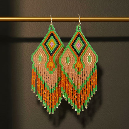 Handgewebte Miyuki Perlenohrringe Modell Tramonto Etrusco von EDELATELIER., dekorativ hängend an einer horizontalen goldenen Metallstange. Das geometrische Muster in Blattgrün, Terrakotta und Mauve sowie die feinen Fransen betonen die filigrane Handarbeit und die Länge von 8,5 cm.