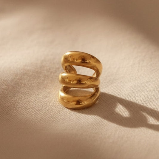 Goldener Ear Cuff ohne Ohrloch mit geschwungener dreifacher Form auf beigem Leinenhintergrund mit weichem Licht von der Seite. 