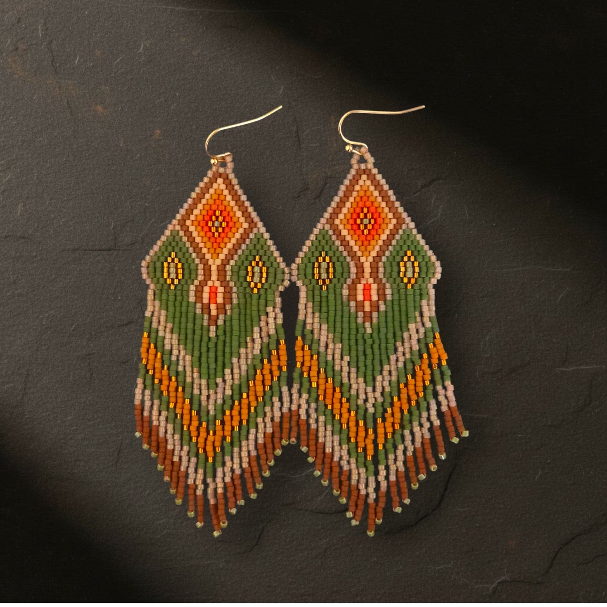 Handgewebte Perlenohrringe Verde Tropicale von EDELATELIER, liegend auf dunklem Naturschiefer. Das Zusammenspiel aus exotischen Grün- und Orangetönen kommt auf dem rustikalen Steinuntergrund besonders kontrastreich und edel zur Geltung
