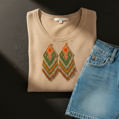 Ein professionelles Flatlay-Arrangement auf dunklem Grund. Die handgefertigten Perlenohrringe in Grün, Orange und Gold liegen auf einem beigen Rundhals-Strickpullover. Rechts daneben ist eine hellblaue Jeans-Shorts platziert. Ein diagonaler Schattenwurf verleiht der Szene eine hochwertige, sonnige Atmosphäre.