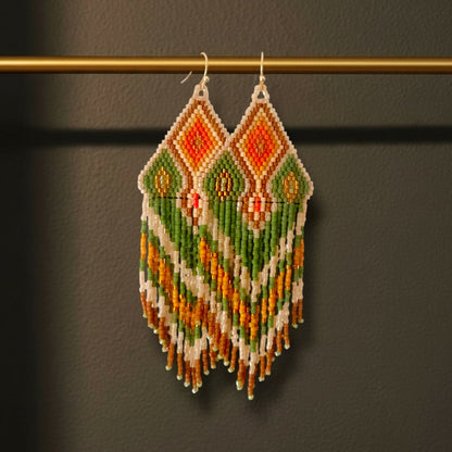 Ein Paar handgefertigte Statement-Ohrringe Modell Verde Tropicale von EDELATELIER, präsentiert an einer goldenen Stange. Die 10 cm langen Ohrringe zeigen ein lebendiges, tropisches Muster in sattem Grün und leuchtendem Orange, kombiniert mit cremeweißen Akzenten und feinen Golddetails.