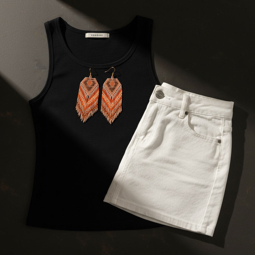 Eine stilvolle Nahaufnahme eines Paares handgefertigter Perlenohrringe von EDELATELIER., platziert auf einem schwarzen Tanktop, neben einem kurzen weißen Jeansrock. Die Ohrringe weisen ein geometrisches Muster in warmen Gelbtönen, Orange und Creme auf und sind mit feinen Perlenfransen versehen. Ein weicher Lichtstrahl fällt auf die Szene und erzeugt tiefe Schatten.