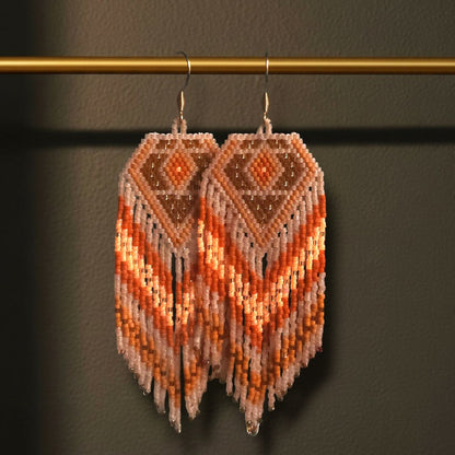 Handgewebte Perlenohrringe Zafferano an einer goldenen Präsentationsstange vor einem dunklen Hintergrund. Das Design zeigt ein warmes Rautenmuster in Braun, Terrakotta und Orange mit langen, fließenden Perlenfransen.